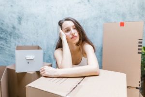 A sad young womne surrounded with cardboard boxes and thinking about hiring professionals when moving house.